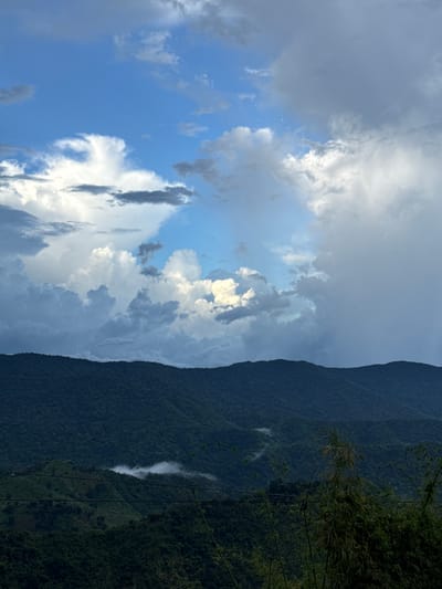 The image depicts a mountainous landscape under a dramatic sky. The foreground consists of dark green, densely forested hills with visible terraced sections on the slopes. A few thin, dark wires are stretched across the middle ground. In the distance, the undulating peaks of more, darker green mountains recede into the background. A light mist or fog clings to the valleys between the mountains. The sky is a dynamic mix of deep blue and a variety of clouds, ranging from bright white, puffy cumulus to darker, more ominous gray formations. The lighting suggests it may be late afternoon or early evening, with the sun breaking through some of the cloud cover. The scene is devoid of any visible people or animals. The location is stated as Nao Sẻ, Vietnam.