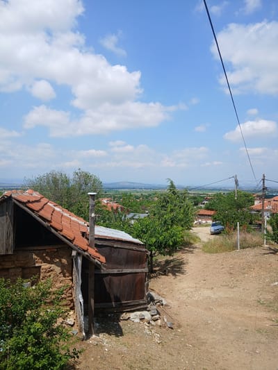 The image depicts a bright, sunny rural landscape, rotated 90 degrees counter-clockwise from its natural orientation. The scene, located in Vetren Dol, Bulgaria, is bathed in a clear blue sky with scattered white cumulus clouds, and strong shadows suggest midday or early afternoon.
In the lower foreground (when viewed correctly), an old, rustic building or shed with a weathered red-tiled roof and exposed brick or mud-built walls is visible. Next to it stands a large, possibly rusted metal barrel or tank, surrounded by stones and rubble. Vibrant green bushes and small trees grow around these structures.
A dusty, unpaved road or path cuts through the midground, sloping gently from right to left. A silver car is parked near the road on the right. Several utility poles with overhead power lines crisscross the upper part of the frame, extending into the distance.
The background opens up to a vast valley or plain, featuring scattered houses and fields, with distant, hazy hills or mountains forming the horizon. The scene is quiet, showing no explicit human activity. No text is visible within the image.