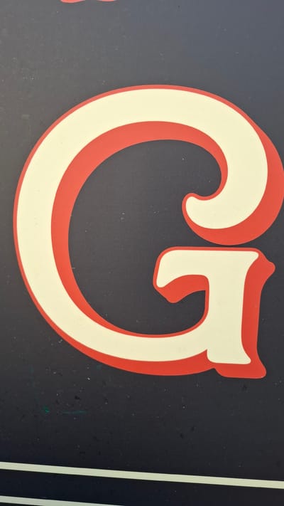 The image is a close-up photograph of a large, stylized letter "G" against a dark, textured background. The "G" is outlined in red and filled with a cream or off-white color, giving it a bold and graphic appearance. The red outline has a rounded, almost three-dimensional quality. The dark background appears to be a flat surface, possibly a wall or a sign, with some subtle speckling or texture visible. There are two thin, light-colored horizontal lines at the very bottom of the image. No specific location in Plovdiv, Bulgaria, can be identified from this image, as it focuses solely on the letterform and its immediate surroundings.