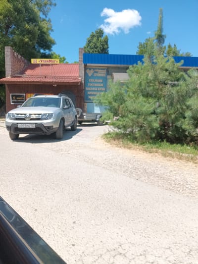 A silver Dacia Duster SUV with license plate HO91MA193 is parked on a paved road. Behind the SUV is a small, light-colored utility trailer. To the left of the SUV, a building with a red tiled roof and a brown facade has a yellow sign that reads "УпакМаг". To the right of the SUV is another building with a blue corrugated metal roof and a light-colored facade. A tall, green pine tree stands in front of the right-hand building. The sky is blue with a single white cloud. The scene appears to be in an outdoor commercial area in Krymsk, Russia.