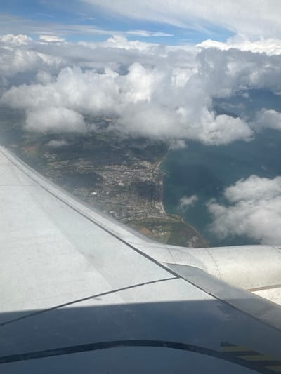 An aerial view shows a coastline in Dublin, Ireland, with the wing of an airplane in the foreground. The wing, made of white metal panels with visible rivets, dominates the lower portion of the image. A dark shadow from the wing falls across its surface.
Beyond the wing, a city sprawls along the edge of the sea. The urban area features a dense collection of buildings, with a clear division between developed land and green spaces. A road network is visible, winding through the city and towards the coast. The sea itself is a deep blue-green, with some whitecaps indicating choppiness.
The sky above is a patchwork of blue sky and fluffy white clouds. The clouds are dense in some areas, obscuring parts of the land below, while in others, they are more scattered, allowing clear views of the city and coastline. The overall impression is of a bright, clear day from a high vantage point.