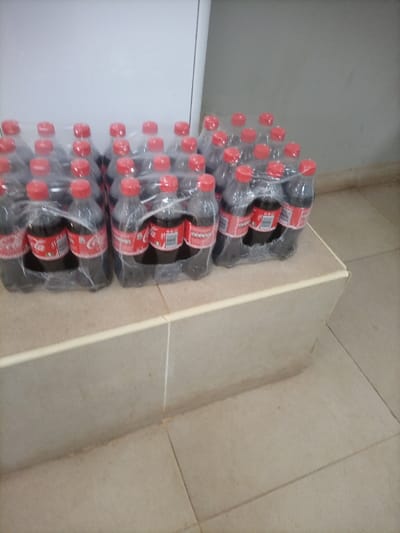 Several plastic-wrapped six-packs of Coca-Cola bottles rest on a ledge covered in tan tiles. The bottles are all red-capped and appear to be full. The ledge is tiled with what looks like ceramic or porcelain. Behind the ledge, a white door or cabinet is visible, and the wall behind the ledge is painted a neutral color. The floor, also tiled in tan, stretches to the lower right corner of the image.