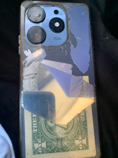 The image shows the back of a cell phone with a protective case. The case appears to be damaged and peeling in several places, with a one-dollar bill tucked underneath the case. The phone has a triple camera system. The image is taken from a close-up perspective, focusing on the back of the phone. No specific location in El Valle del Espíritu Santo, Venezuela, can be determined from the image.