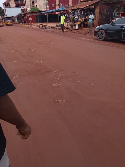 This image depicts a street scene in Enugu, Nigeria, characterized by a dusty red dirt road. In the background, a row of modest market stalls with corrugated metal roofs lines the street. To the left, a modern multi-story building with red and white facades stands, suggesting a mix of development. Several small vehicles, including a yellow auto-rickshaw, are parked or moving on the road.
In the middle ground, two men are visible near the stalls. One man, wearing a bright yellow t-shirt and dark shorts, stands on the left. Another man, in a light blue t-shirt with yellow sleeves and grey trousers, stands to the right, looking at his phone. A black sedan is parked on the right side of the road, partially obscuring the market stalls. The foreground shows a portion of the red dirt road with scattered debris and some lighter-colored patches, possibly indicating areas of hardened soil or loose gravel. The overall impression is of a busy, informal marketplace in a developing urban area.