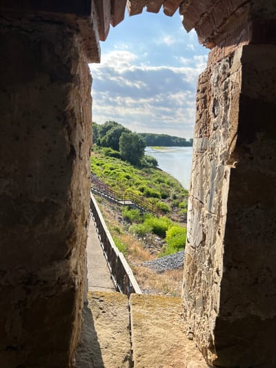 This is an eye-level, vertical outdoor photograph taken through a narrow opening in an old stone structure, possibly a castle or fortification. The opening frames a view of a river, green landscape, and a cloudy sky.
The immediate foreground consists of rough, textured stone in shades of brown and grey, with visible bricks on the left side of the frame. These stones form the walls of the structure through which the photo is taken, creating a sense of looking out from within.
Beyond the stone opening, the scene opens up to a river in the distance, bordered by a lush green hillside densely covered with trees and vegetation. A pathway or set of stairs with a metal railing is visible ascending the hillside from the riverbank, suggesting a recreational area or access point. The sky is partly cloudy with patches of blue, indicating daylight, likely late morning or early afternoon given the light and shadows.
The overall impression is one of historical architecture juxtaposed with a natural landscape. There is no indication of people or any specific activity beyond the static scenery. The location context suggests this might be a view from a historical site in Belene, Bulgaria, overlooking the Danube River. The emotions evoked are contemplative and peaceful, with a sense of history and the natural world coexisting. No visible text is present in the image.