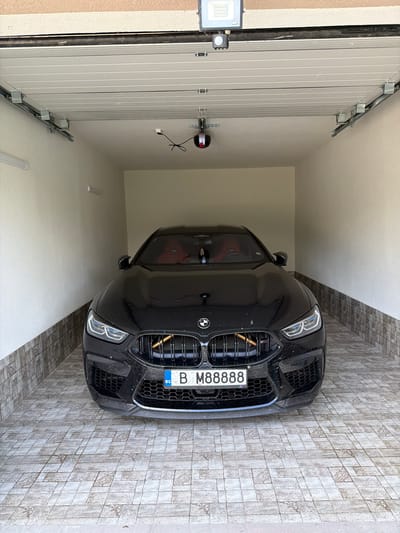 A black BMW M8 Coupe is parked centrally inside a well-lit garage, facing the viewer. The vehicle's front bumper, finished with carbon fiber-look trim, appears slightly dusty. Its large, black twin-kidney grille features vertical slats, behind which a golden or bronze stick-like object is horizontally positioned. Modern LED headlights with distinct daytime running lights are illuminated. The interior, visible through the windshield, showcases prominent red leather seats. The front license plate displays "B M88888" and includes the "BG" (Bulgaria) country code identifier.
The garage features smooth, light-colored walls. The lower sections of the walls are tiled with brown and gray rectangular tiles arranged horizontally. The floor is covered with light gray and beige square ceramic tiles, some of which exhibit subtle decorative patterns. Overhead, a sectional garage door, composed of light-colored horizontal panels, is in a partially open position, revealing its metal tracks and an automated garage door opener motor (black and red) mounted to the ceiling. A rectangular LED floodlight (silver/gray) is mounted above the garage entrance, just outside the opening. Two tubular LED light fixtures are mounted horizontally on the upper left wall, with another partially visible on the right wall.
This scene is located in Targovishte, Bulgaria.