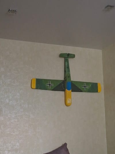 A wooden model of a German World War II aircraft is mounted on a light-colored wall. The model is painted with green camouflage, yellow highlights on the fuselage and wingtips, and black crosses. The model is horizontally oriented with the tail on the left side of the image. The wall behind the model is textured. The location is the city of Serpukhov, Russia.