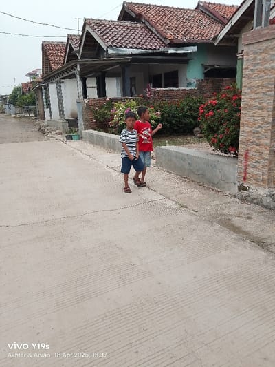 The image shows two young boys standing on a paved road in Anjatan, Indonesia, located at latitude -6.412 and longitude 107.962. The road is lined with houses, some of which have red tile roofs. There are bushes with red flowers and a brick wall in the background. The boy on the left is wearing a black and white striped shirt and blue shorts, while the boy on the right is wearing a red shirt and blue shorts. The boys are looking at something in the distance.  The date and time are stamped on the bottom right of the image: "18 Apr 2025, 13.37."