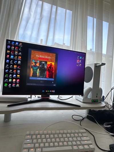 The image shows a desk with a computer monitor, a white Xbox Series S, and a white mechanical keyboard. The monitor displays the Steam store page for "The Elder Scrolls" franchise. The desk is positioned near a window covered with white blinds. The environment appears to be a home office or gaming setup. The image provides no discernible information connecting it to Sevlievo, Bulgaria.