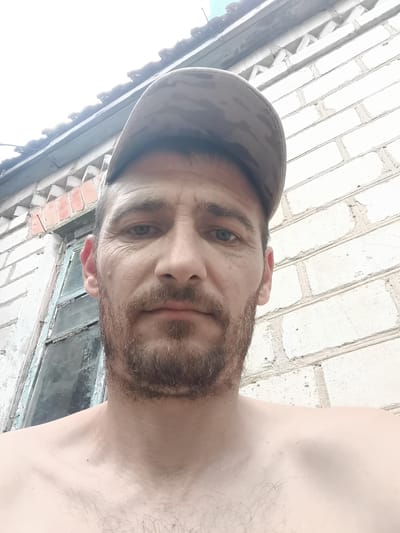 The media file is a close-up, low-angle selfie of a shirtless man wearing a camouflage baseball cap. He has short, brown hair and a light beard. His eyes are blue and he has a neutral expression. The background shows a white brick wall with some signs of wear and tear, and a portion of an old, weathered window with peeling blue paint. Above the wall, a section of a roof is visible, with some sunlight filtering through. The overall setting appears to be outdoors, possibly in a rural or older urban area. The lighting suggests it is daytime, with bright, overcast conditions. There are no visible text elements in the image. The location context provided is Saksahan, Ukraine.
