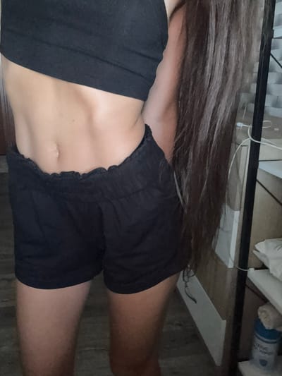 The image shows a close-up of a person's torso and legs, wearing a black crop top and black shorts. The person has long, dark brown hair that extends down their right side. Their midsection is visible, showing toned abdominal muscles. The background appears to be an indoor setting, possibly a room, with a shelf and some items on it, including a blue container. There are no visible indications of the specific location within Basarabeasca, Moldova.