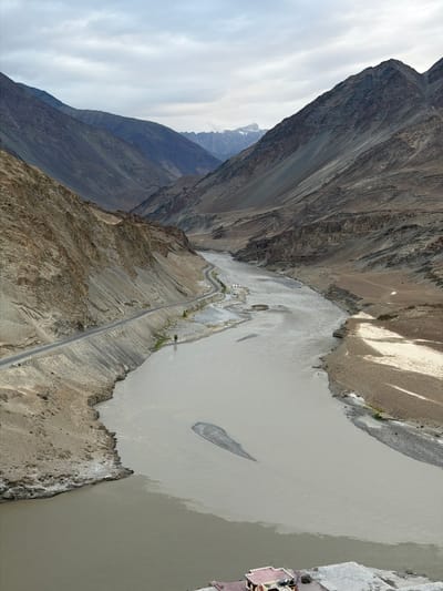 A high-angle, vertical shot captures a wide, shallow river flowing through a rugged, mountainous valley. The river's water is a milky, light brown, with darker, more turbulent currents visible where two bodies of water appear to merge. On the left bank, a winding paved road hugs the steep, rocky slope, which is covered in loose scree and sparse vegetation. A small, white building with a red roof is visible at the bottom right, situated on a paved area near the riverbank. The valley walls are steep and barren, showing layers of sedimentary rock formations. In the distance, the valley narrows, revealing more rugged peaks under a cloudy, overcast sky. The scene depicts a remote, natural landscape with minimal human presence, suggesting a location like the confluence of the Indus and Zanskar rivers near Leh, Ladakh, India, an area known for its dramatic mountain scenery and glacial rivers.
