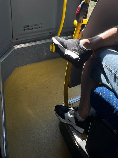 The image shows a close-up of two pairs of feet wearing athletic shoes inside a vehicle, likely a bus or train. The perspective is from above, looking down. One person is wearing black and white New Balance sneakers with white socks, and the other person is wearing black and white sneakers with white socks. The shoes are placed on a yellow floor with some dark speckling.

The setting appears to be public transportation, with a yellow handrail visible in the foreground and a blue seat with a pattern of colorful dots in the background. A portion of denim shorts is visible, suggesting the person is sitting. Sunlight is casting strong shadows, indicating it's daytime, likely sunny. A red button with the word "STOP" is partially visible on the yellow handrail. The overall impression is of a mundane moment of travel. The location context is Dobřany, Czechia.