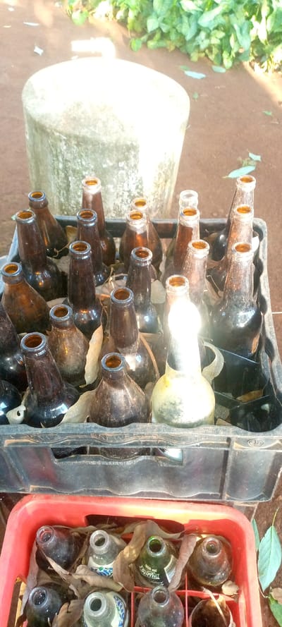 Two crates of empty bottles sit outdoors. The top crate is black and contains brown beer bottles, interspersed with dry leaves. One bottle is light-colored, reflecting the bright sunlight. Below, a red crate holds more empty bottles, including some with labels like "Sprite" visible. Dry leaves also fill this crate, obscuring the bottles. In the background, a concrete stool and some green plants are visible on the brown earth.
