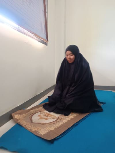 A woman in a black hijab and robe is seated on a tan and brown prayer rug layered on a blue mat. She is positioned near a white wall and appears to be in prayer. A framed document or chart is affixed to the wall behind and to the left of her. The scene is lit by natural light from an unseen window.