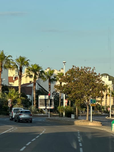 This is a photograph taken from a street in Toulon, France, looking towards buildings and a road with palm trees lining the side. The sky is clear and blue with a few faint clouds. In the distance, white buildings with red-tiled roofs are visible, interspersed with tall palm trees. A traffic light is visible on the right side of the frame, displaying a red light. Two cars are driving on the road, a silver SUV and a white van behind it. A road sign indicating "A57 HYERES NICE" is also present. The scene appears to be a sunny day in a Mediterranean coastal city.