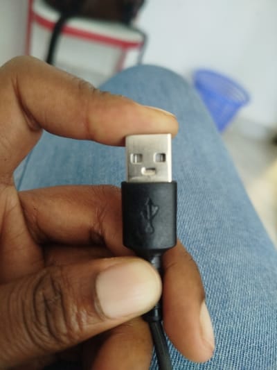 A close-up photograph shows a dark-skinned human hand holding a black USB-A male connector. The hand's thumb is positioned on top of the connector's metallic end, while the index finger supports the black plastic housing from below. The silver-colored, rectangular metal tip of the USB connector is oriented towards the viewer, displaying its four internal rectangular pin openings. Below the metallic tip, the black molded plastic portion of the connector features a raised USB trident logo. A black, ribbed cable extends downwards from the connector. The background immediately behind the hand consists of blue denim fabric, likely part of trousers. In the blurred far background, a white wall or surface is visible, along with a partial red and white striped object on the upper left, and a partial blue plastic container on the upper right. The scene is located in Keekonyokie ward, Kenya.