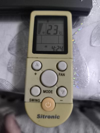 This is a close-up, eye-level shot of a beige air conditioner remote control with a digital display. The remote is positioned vertically in the center of the frame, resting on a textured, light-colored surface that appears to be fabric, possibly a tablecloth or sofa upholstery, with a patterned design. The background is blurred, showing more of the same fabric and dark, indistinct shapes above.
The digital display on the remote shows "23°C" at the top, indicating the current temperature. Below that, it displays "AM 4:24", suggesting the time. The remote has several buttons: a circular power button with a white circular icon, a larger central button labeled "MODE", and other smaller buttons arranged around it, including directional arrows (up and down triangles), a plus sign, and a button labeled "FAN". There's also a button labeled "SWING". The brand name "Sitronic" is visible in dark text at the bottom of the remote. The overall lighting is soft, and there are no explicit indications of weather or specific emotions. The context of Kokand, Uzbekistan, is provided but not visually discernible from the image itself. The focus is entirely on the remote control.