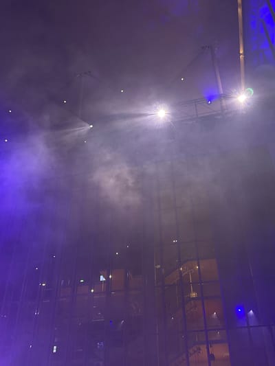 The image depicts an indoor scene with dramatic lighting and atmospheric effects, likely a concert or theatrical performance. The primary subjects are beams of light cutting through a hazy or smoky atmosphere, illuminating a stage structure. Several bright spotlights are visible, casting sharp rays downwards. The background features a large, multi-paned glass structure, suggesting a modern building or arena. Reflections of interior lights and what appear to be staircases are visible within the glass, hinting at balconies or upper levels. The overall color palette is dominated by purples and blues from the stage lighting, with some white light from the spotlights. The presence of fog or haze is a notable detail, creating a theatrical ambiance. There are no discernible people in the image, but the setup clearly indicates a performance setting. The time of day is not directly indicated, but the artificial lighting suggests it could be evening or night. The location is specified as Athens, Greece, but no specific landmarks are visible to confirm this. There is no visible text in the image.