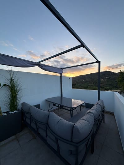 This image captures an outdoor patio or balcony area in Loulé, Portugal, during what appears to be sunset or dusk. The scene is dominated by a modern, dark grey outdoor seating set, including a sectional sofa with plush cushions and a matching rectangular coffee table. A dark grey metal pergola structure with a retractable canopy extends over the seating area. To the left, a tall, dark planter contains a long-bladed green plant, possibly ornamental grass, accented with white stones. The patio floor is tiled with large, grey square tiles. Beyond the patio, a low white wall encloses the space, revealing a hilly landscape with sparse vegetation and distant, small white structures, illuminated by the warm hues of the setting sun. The sky is a gradient of soft blue with streaks of orange and pink clouds, suggesting the transition from day to night. No people are present in the image.