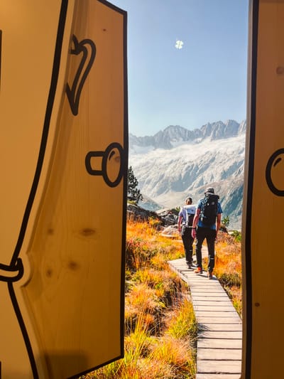 The image portrays an indoor display or large print designed to simulate an open wooden door revealing a mountain landscape. The foreground features two vertical, light-toned wooden panels acting as an open doorway, each adorned with thick black cartoon-style outlines and simple black graphic elements, including a heart-like shape on the left panel and a circular element on both.
Through this simulated opening, a high-alpine environment is depicted under a clear blue sky, containing a faint, blurry white four-leaf clover shape near the top center. The background is dominated by rugged, snow-capped or glaciated mountains with rocky peaks and extensive ice formations. The midground and foreground of the framed scene present an autumnal landscape, characterized by dense, vibrant orange, yellow, and reddish-brown grasses and low vegetation over rocky terrain. Scattered dark green coniferous trees are visible on the left side, and a body of water is faintly discernible in the mid-distance on the left.
A wooden boardwalk path, composed of horizontal planks, extends from the lower center of the framed scene, curving slightly to the left and receding into the landscape. Two individuals are walking in single file away from the viewer on this boardwalk. The person closer to the foreground, on the right side of the path, wears dark clothing, a large dark hiking backpack, and bright orange shoes. The person ahead, on the left, wears lighter-colored clothing and a lighter-colored hiking backpack. Both are equipped for mountain trekking.
This display is situated in Erstfeld, Switzerland, showcasing a typical Swiss mountain vista.