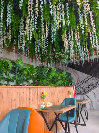 This image captures an interior view of a vibrant and inviting dining or lounge area in Semenovka, Kyrgyzstan. The ceiling is densely adorned with artificial green foliage and numerous cascading strands of white and pale pink wisteria-like flowers, creating an overhead canopy that dominates the upper portion of the frame. Beneath this floral ceiling, a long wooden partition separates the foreground seating from a lush display of artificial palm plants, adding a natural and tropical feel to the space.

In the foreground and midground, several modern dining tables and chairs are arranged. The chairs feature striking teal-blue and orange upholstery, providing pops of color against the natural wood tones of the tables and partition. The tables are light-colored wood with dark, angular metal bases, each set with dark green placemats. Decorative elements on the tables include small vases with artificial flowers – red and yellow tulips on one, and pink blooms with a sugar bowl on another. A small wooden box on one table bears the legible text "ALTIN KAZYK," and a tag with the number "20" is visible on another table.

To the right, an elegant dark metal staircase with an ornate railing ascends against a wall featuring light-colored, marble-effect panels and dark brickwork. No people are visible, indicating the space is unoccupied. The overall ambiance is one of calm and pleasantness, with a blend of natural and contemporary design elements. The lighting appears bright and uniform, typical of an indoor commercial establishment.