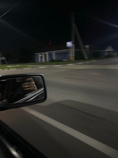 The image captures a nighttime scene from the perspective of someone in a vehicle, looking out the side window and at the side-view mirror. The primary subjects are the reflection of a person's hand in the side-view mirror and the blurred motion of the outside environment.
In the side-view mirror, a hand is visible, seemingly resting or reaching towards the camera. The rest of the reflection is dark and indistinct, suggesting the interior of the vehicle. Outside the vehicle, the scene is greatly blurred due to motion, but discernible elements include a road with lane markings, streetlights, and buildings. A blue and white sign is visible on a pole in the distance. To the far left, there are red lights, possibly on a building or structure. The overall lighting suggests it is nighttime. The setting appears to be an urban or semi-urban area, consistent with a location in Kostanay, Kazakhstan. The motion blur indicates the vehicle is in transit. There is no visible text in the image.