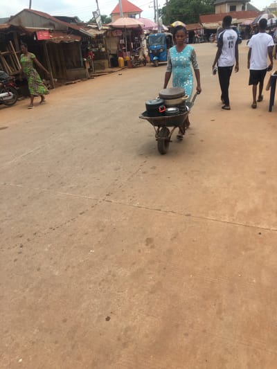 The image depicts a street scene in Enyibichiri, Nigeria. A woman wearing a blue dress walks along a concrete road, pushing a wheelbarrow loaded with metal pots and pans. Two young men wearing white shirts walk in the same direction, parallel to the woman. In the background, there is a blue "Keke Napep" (tricycle taxi) parked near stalls and shops. One stall has a variety of shoes displayed. A second woman in a green patterned dress is standing next to a store. A motorcycle is parked next to the store.