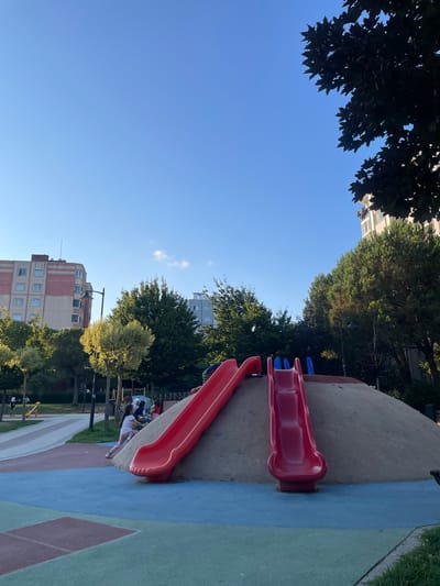 The image depicts a public park with a playground in Başakşehir, Turkey. The foreground features a sand-covered mound with two vibrant red slides. One slide is longer and curves to the left, while the other is shorter and straighter, descending to the right. A young girl in a pink shirt and white pants is actively playing on the mound, partially obscured by the sand. In the mid-ground, several trees with lush green foliage are visible, interspersed with buildings. One building on the left is tall with a pink and beige facade. Another building on the right is lighter in color and also appears to be residential. The sky above is a clear, bright blue, indicating a sunny day. The ground of the playground is a mix of blue, green, and red patterned surfacing. In the background, there are more trees and hints of urban infrastructure. The scene captures a moment of outdoor recreation in a city park environment.
