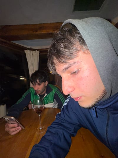 Two men are captured in a dimly lit interior. One, wearing a grey beanie and a dark blue tracksuit, appears closer to the camera. Below him, a second man is visible in the image, reflected in the glass of a table. He's wearing a green and blue tracksuit, holding a cell phone in his hand. A wine glass is situated between them. The image is captured at an awkward angle, with the subjects seemingly tilted. A window with drawn white curtains is visible in the background.