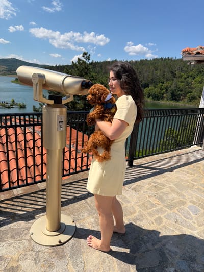 In the city of Banya, Bulgaria, a young woman stands barefoot on a stone patio, holding a small, fluffy brown dog. She is wearing a light yellow dress. The woman is looking at a large, gold-colored coin-operated telescope positioned to her left. The telescope is pointed towards a scenic view of a calm lake, surrounded by lush green trees and rolling hills under a bright blue sky with scattered white clouds. A black metal railing runs along the patio edge, overlooking the lake. The patio is paved with irregular grey and brown stones, and shadows from the railing and a nearby structure are cast across it. In the background, a building with a red-tiled roof is visible on a hill. The overall scene is one of leisure and enjoyment of the natural landscape.