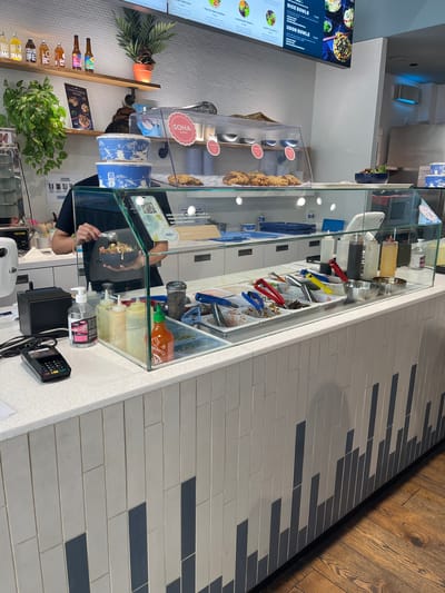 The image shows the interior of a restaurant called "Soha" in Lyon, France. A person is preparing a meal behind a glass counter displaying various ingredients and sauces. The menu board on the left features "Rice Bowls" and "Udon Bowls." A POS system is on the counter. Shelves behind the counter display plants and drinks. The interior features a brick and tile design.