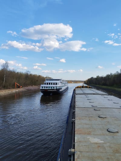 The image shows a canal in Varvarino, Russia. A multi-decked river cruise ship passes a large barge loaded with concrete panels. On the left bank, an orange excavator sits along the edge of the trees. The sky is blue with scattered white clouds.