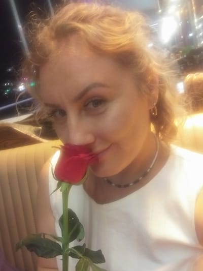 The image is a close-up, selfie-style shot of a woman holding a red rose to her nose. She has fair skin, blonde hair, and appears to be wearing a white top and a beaded necklace. The background is blurred but suggests an indoor setting with warm lighting and some out-of-focus lights that could be from a city environment. The woman is positioned in the foreground, with the rose obscuring her mouth. The overall impression is intimate and romantic. The image does not contain specific visual cues that would allow for precise determination of the location being Beyoğlu, Turkey.
