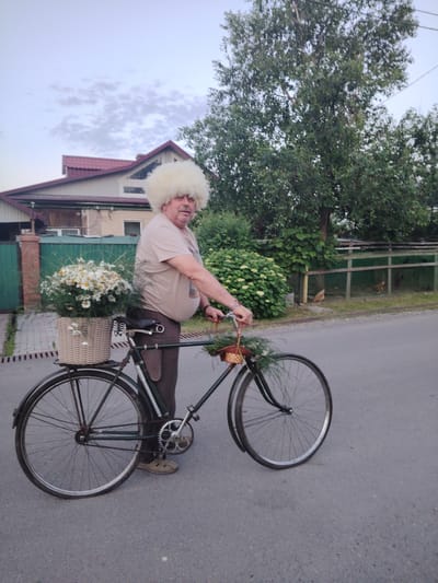 A man with a large, fluffy white wig is standing next to a bicycle, holding onto the handlebars. He is wearing a light brown t-shirt and dark pants, with light brown shoes. The bicycle has a woven basket filled with daisies attached to the rear rack. A smaller woven basket with greenery is attached to the front handlebars. The man has a pot-bellied physique and appears to be middle-aged or older.
The scene is set outdoors on a paved road, with a residential building and trees in the background. The building has a red tiled roof and light-colored walls. A green fence separates the property from the road. Behind the fence, there is some greenery and two chickens can be seen in an enclosure. The sky is overcast with soft clouds, suggesting it is either early morning or late afternoon.
The overall mood is relaxed and perhaps a bit whimsical, given the man's unusual wig. The presence of the flowers and the bicycle suggests a leisurely activity, possibly a ride or preparation for one. The location context of Ussurijsk, Russia, provides geographical information for the scene. There is no visible text in the image.