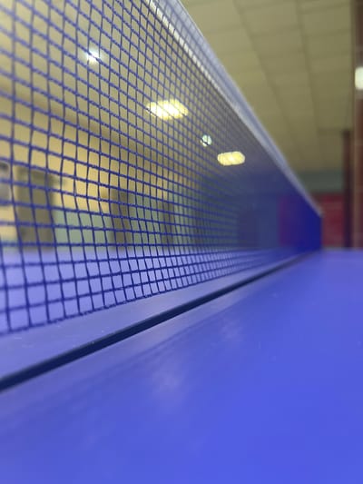This is a close-up, abstract photograph that focuses on the texture and color of materials. The dominant subject is a blue surface on the left, which appears to be a flat, smooth panel. To the right of this panel, a fine blue mesh grid is visible, creating a textured overlay. Through the mesh, a background is discernible, featuring a blurry, light-colored surface that could be a wall or ceiling, with several bright, elongated yellow lights reflected or embedded within it. The composition is abstract due to the close-up perspective and the lack of clear context for the objects.
The image does not depict any people or a specific activity. It is likely an indoor scene, given the presence of artificial lighting. The bright lights suggest it could be daytime or nighttime, as artificial illumination is present. There are no overt location cues within the image itself, but the provided context places it in Satbayev, Kazakhstan. The overall mood is neutral and objective, focusing on the visual properties of the materials. No text is visible in the image.