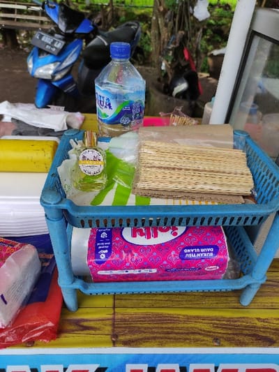 The scene is a street food vendor stall in Kecamatan Lawang, Indonesia (latitude: -7.834, longitude: 112.693). The vendor is selling food items from a blue metal shelf. The shelf is positioned on a wooden table. The shelf contains a box of tissue paper, a stack of wooden skewers, and a bottle of clear liquid. A blue scooter is visible in the background, partially obscured by vegetation. There is also a partially obscured yellow object on the table in the foreground. The environment is outdoors, likely on a street or sidewalk.
