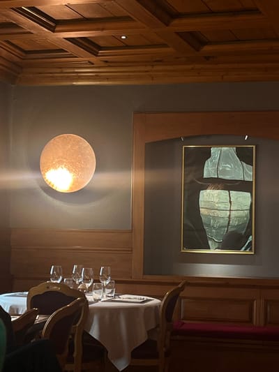 The image depicts the interior of a dining establishment, identified as the Restaurant du Parc within the Hôtel de l'Ill in Sélestat, France.

The scene is characterized by warm, subdued lighting and rich wooden accents. The ceiling is a detailed coffered design constructed from light brown wood, featuring multiple square and rectangular panels, with recessed lighting points. The upper sections of the walls are painted a solid medium gray. Below this, approximately waist-height, runs a consistent wooden wainscoting that extends across the visible walls, matching the ceiling's wood tone. Vertical wooden frames and an arched wooden alcove on the right further segment the wall space.

On the left wall, a prominent circular wall sconce, textured and glowing with a warm, bright light, casts a noticeable glare. On the right wall, set within the arched wooden alcove, hangs a rectangular artwork framed in gold. The artwork itself is abstract, displaying a segmented form in muted light green and blue tones against a dark background, possibly representing a draped figure or torso. It is illuminated by two small spotlights mounted above the frame.

In the mid-ground, a dining table covered with a white tablecloth is visible, set with multiple stemmed wine glasses, water tumblers, white plates, and cutlery, suggesting readiness for service. Partially visible to the left of this table is another table, also draped in a white tablecloth. Dining chairs with wooden frames and dark, possibly red or purple, upholstered backs are positioned around the tables. Along the wall on the right, beneath the framed artwork, is a built-in wooden bench with a red upholstered cushion.

A portion of an individual, dressed in a dark garment, is partially visible in the bottom-left corner of the frame, indicating the presence of a person seated out of view. No explicit actions or interactions are discernible.