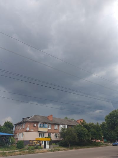 In the city of Hadiach, Ukraine, a brick apartment building stands under a dark, cloudy sky. The building has multiple windows, some with blue shutters, and a gray shingled roof. Power lines crisscross the sky above the building. In front of the building, there is a small business with a yellow awning and a sign that reads "ФОТО НА ПОЛОТИ" which translates to "Photo on the screen." A woman is standing near the entrance of the business. Trees and greenery surround the building, and a road runs in front of it. A blue car is parked on the right side of the road. The overall scene suggests an overcast day, possibly before or after a rainstorm.