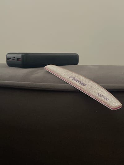 A black rectangular power bank is positioned on a gray upholstered surface. To the right of the power bank, a pink and white emery board lies diagonally across the surface. The emery board has the text "WAY SKO" and "100/180" printed on it. The background consists of a plain, light-colored wall. The scene is indoors. The location of this image is not specified in the city of Kommunarka, Russia.