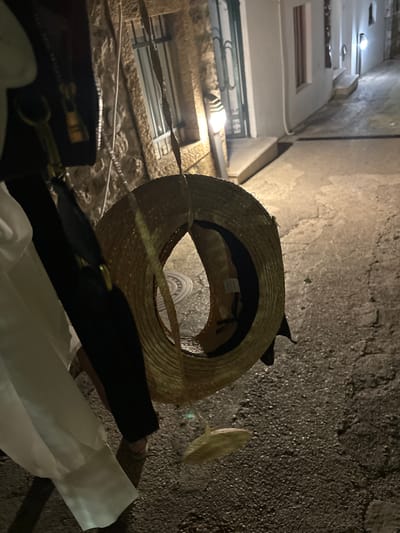 In Petrovac centar, Montenegro, a close-up shot focuses on a straw hat dangling from a dark purse strap, held by an unseen person. The purse, partially visible on the left, has a gold zipper and a looped pull. The hat is woven straw with a dark band and a label visible inside. Behind the hat and purse, a textured stone wall with a barred window is dimly lit. To the right, a narrow, paved alleyway is illuminated by a wall-mounted lamp, casting a warm glow on the damp asphalt. The scene appears to be set at night, with the alley stretching into the background.