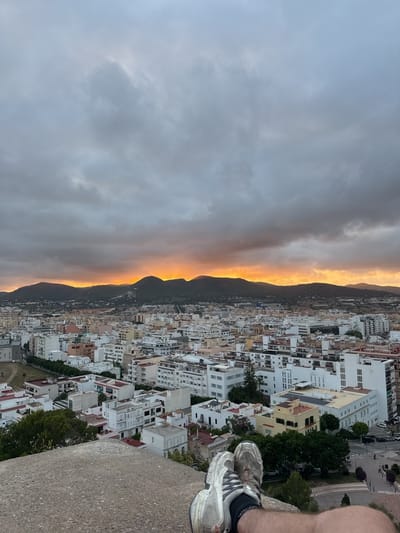 The image presents an elevated panorama of Eivissa (Ibiza Town), Spain, captured from an elevated position likely within Dalt Vila, overlooking the urban area and surrounding landscape. In the immediate foreground, the lower extremities of an individual, clad in dark socks and light-colored athletic shoes, are visible, resting on a rough, textured concrete or stone ledge. The shoes appear worn and dusty.
Below the ledge, the cityscape of Eivissa extends across the midground. The urban area is characterized by a dense arrangement of predominantly white and light-colored multi-story buildings, exhibiting modern and Mediterranean architectural styles with flat roofs. Interspersed throughout the built environment are areas of green foliage from trees and shrubs. Roads and smaller pathways are visible weaving through the buildings, and a few parked vehicles are discernible on the right side of the frame.
In the background, a series of dark green, undulating hills or low mountains forms the natural horizon line. Above these hills, the sky is largely overcast with extensive gray and dark gray stratocumulus clouds. A distinct band of vibrant orange and yellow light illuminates the horizon just above the mountains and the lowest layers of clouds, indicating either sunrise or sunset. This warm light contrasts with the otherwise muted, diffused light from the cloud-covered sky.