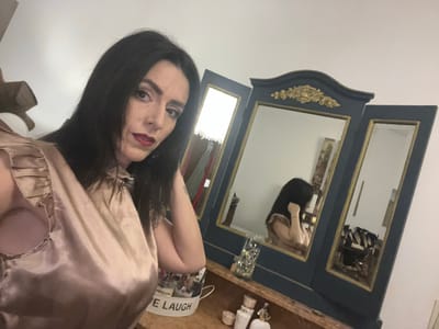 A person with dark hair and red lipstick stands in front of a dark blue dresser with two mirrors. The top of the dresser holds decorative items including a white container labeled "LIVE LAUGH LOVE". The person is wearing a shiny, light-colored garment. The location is unknown.