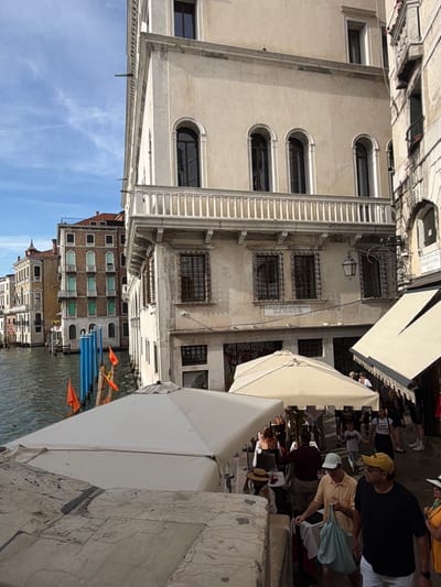 The image depicts a sunlit street scene in Venice, Italy, likely along a canal. The foreground features a stone railing and the white fabric of an awning. In the midground, several people are seen walking and interacting. A man in a yellow hat and black shirt, and another man in an orange shirt carrying a light blue bag are prominent. Behind them, a white umbrella shades a seating area, and a building with arched windows and decorative railings rises in the background. Orange flags are visible near blue poles in the water, suggesting a canal or waterfront area. The architecture is characteristic of Venetian buildings, with multiple stories, traditional windows, and weathered facades. The sky is blue with scattered clouds.