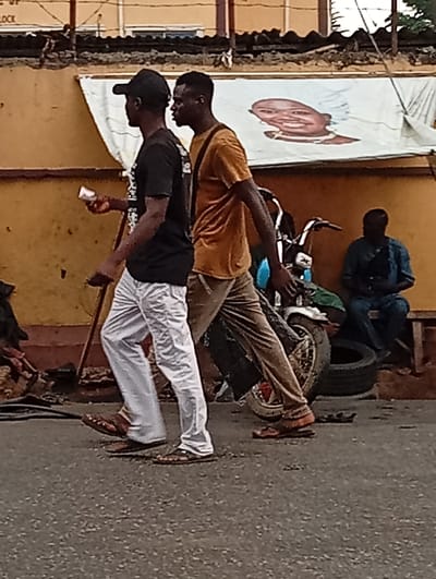 In Shangisha, Nigeria, two men walk down a paved street. The man on the left wears a black t-shirt, a black baseball cap, and white pants. He holds a white object, possibly a cup, in his right hand. The man on the right wears a gold t-shirt and brown pants. A motorcycle with a green seat is parked behind them. To the right of the motorcycle, a man sits on a bench wearing a blue patterned shirt and pants. A large banner with a drawing of a child's face is stretched across a wall behind the men. The wall is painted orange.