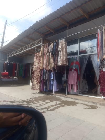 In the city of Razzakov, Kyrgyzstan, an outdoor clothing market stall displays a variety of garments. The stall features a corrugated metal roof supported by wooden beams and a metal truss system. Several racks of clothing hang in front of a storefront with large glass windows. The clothing items include dresses, skirts, and what appear to be two-piece outfits, displayed on mannequins and hangers. A rug is also displayed on a stand. The ground in front of the stall is paved with concrete and pavers, with some wet patches visible, suggesting recent rain or cleaning. A car's side mirror is partially visible in the foreground, reflecting a hand with a ring. The sky above is cloudy.