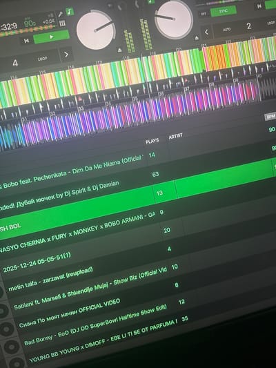 The image shows a close-up, angled view of a DJ setup, likely in a dimly lit room or studio. The primary objects are digital interfaces for music control, including turntables, faders, and waveform displays that are illuminated by colorful LED lights. The scene is dominated by green and dark gray screens displaying song titles, artist names, BPM (beats per minute), and play counts.
Several song titles and artist names are legible, such as "Dim Da Me Niama (Official Video)," "Sabiani ft. Marseli & Shkendije Mujaj - Show Biz (Official Vid," "Bad Bunny - EoO (DJ OD SuperBowl Halftime Show Edit)," and "YOUNG BB YOUNG x DIMOFF - EBE LI TI SE OT PARFUMA." Numerical data like BPM, plays, and track numbers (e.g., 32:9, 90, 14, 63, 13, 9, 20, 4, 10, 6, 12, 35) are also visible.
The activity taking place is music mixing or DJing. The colorful waveforms suggest music is playing or being manipulated. The overall mood is focused and technical, with no people clearly visible in the frame. The lighting is artificial and comes from the equipment itself. The mention of "Sevlievo, Bulgaria" in the prompt does not appear to have any direct visual cues within the image itself, but provides an external context for the location. The image is likely taken at night or in a controlled lighting environment due to the darkness of the surroundings and the prominence of the illuminated screens.