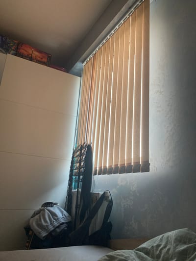 The image depicts an indoor scene, possibly a bedroom, with a window covered by vertical blinds. Sunlight streams through the blinds, casting a pattern of light and shadow on the wall. In the foreground, a pile of clothing is visible, with a wig and a hairbrush on top. A portion of a bed with a patterned duvet is also visible in the lower right corner. The wall to the right has a textured finish with a mottled gray appearance. To the left, a large white cabinet or wardrobe is present, with a jigsaw puzzle box featuring an image of London on its top surface. The scene is likely set in a residential area, and based on the location tag, it is in Karlovo, Bulgaria.