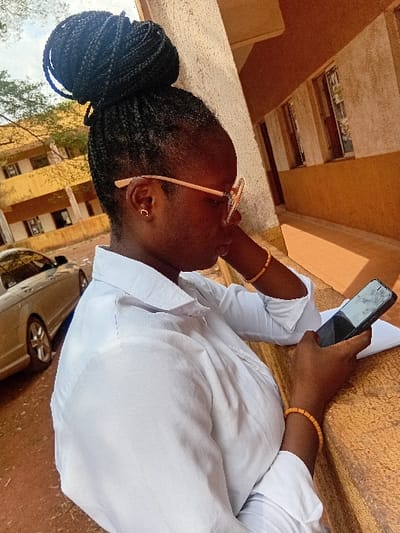 A young, dark-skinned woman with braided hair in a bun wears round glasses. She holds a cell phone in her right hand, while her left arm is bent. A light-colored vehicle is near her. She wears a white shirt and beaded bracelets on both wrists. The scene appears to be outdoors, near a building with yellow-colored walls and brown accents. The photograph is rotated, with the top of the image oriented towards the left.