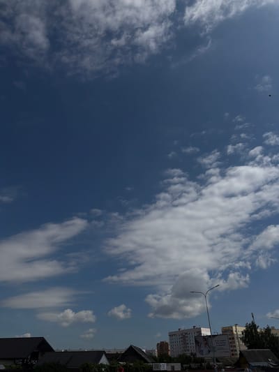 The image shows a wide, upward-facing view of a bright blue sky filled with fluffy white clouds. In the lower portion of the image, a cityscape is visible, featuring the rooftops of several buildings, including some houses with dark, sloped roofs and several multi-story apartment buildings. A streetlamp is prominent in the mid-ground, and a billboard with text and an image is attached to one of the buildings. Some trees and other vegetation are visible between the buildings. A small, dark object, possibly a bird or a drone, is visible against the sky in the upper right quadrant. The overall scene suggests an outdoor environment during daytime, likely in a developed urban or suburban area. The presence of the specific apartment building styles and the Cyrillic text on the billboard point to a location in Belarus. Based on the context of similar images and architectural styles, this scene is likely from Mahilyow, Belarus.